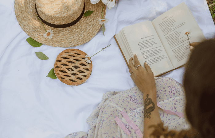 A lady reading outside, she is on a white blanket, there is a straw hat, daisies and fruit pie next to her